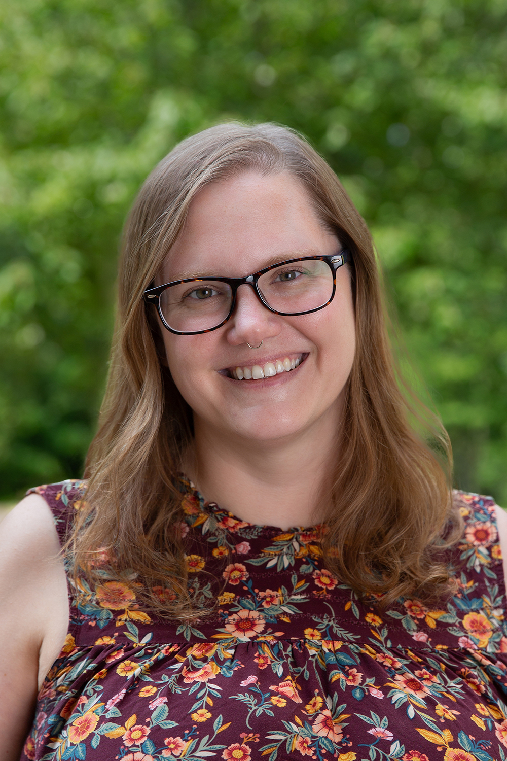 photograph of emily joy zeller, who is smiling and wearing glasses and a brown floral sleeveless shirt